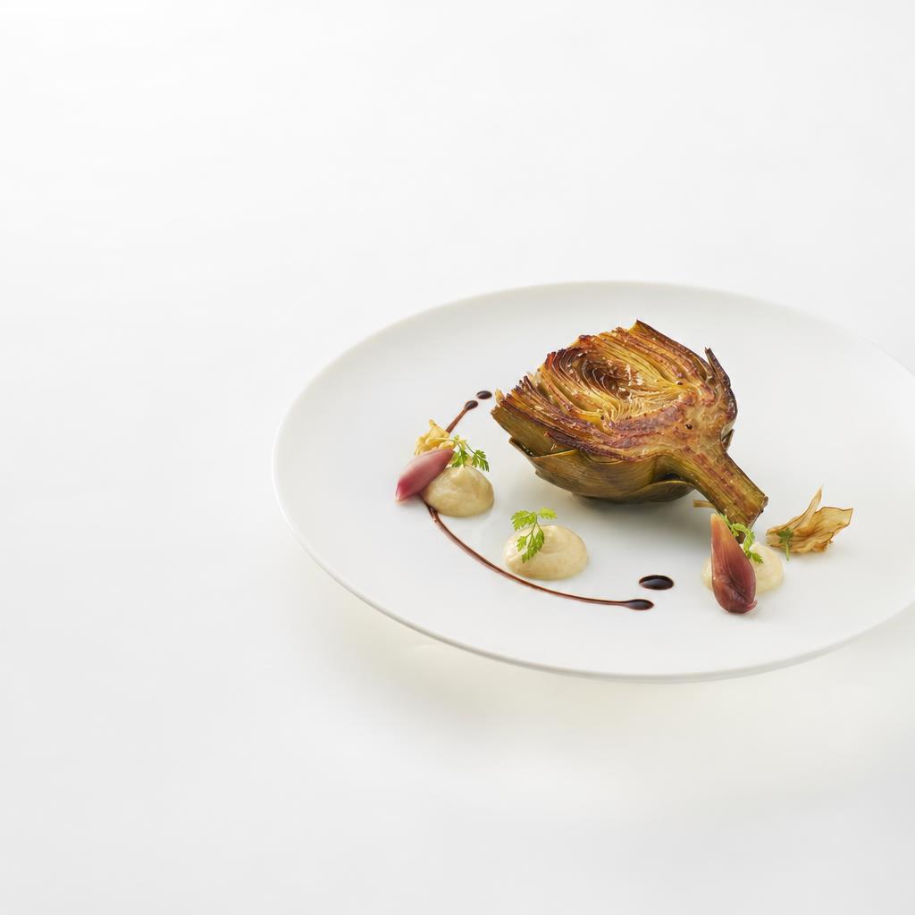 Geröstete Artischocke mit heller Tonkacreme, dunkler Sauce aus schwarzem Knoblauch und eingelegten Magnolienknospen auf weißem Teller.