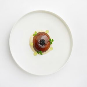 Lackierter Steinpilzhut auf heller Kombu-Tonka-Crème mit Kaviar und kleinen Verjusgel-Punkten auf weißem Teller.